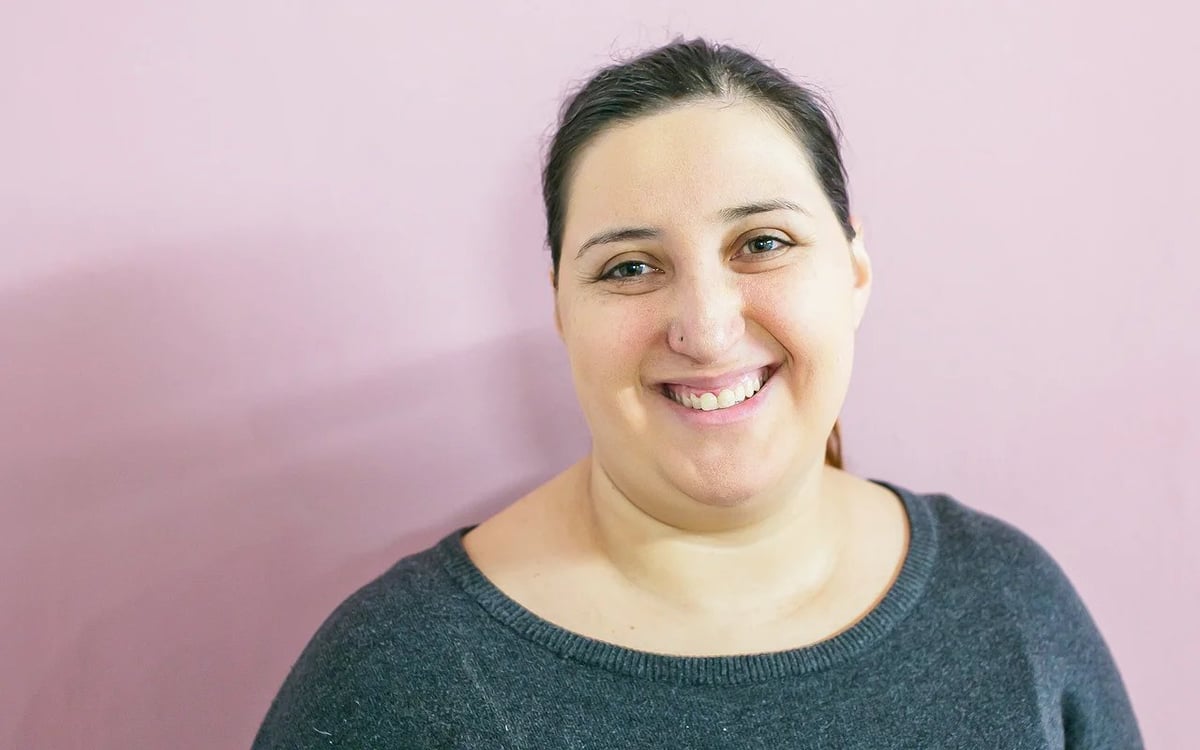 Portrait of a smiling woman with dark hair wearing a dark gray sweater against a pink background