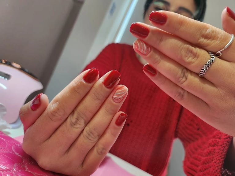 Woman displaying manicured red nail polish with metallic accents and decorative ring against red fabric background